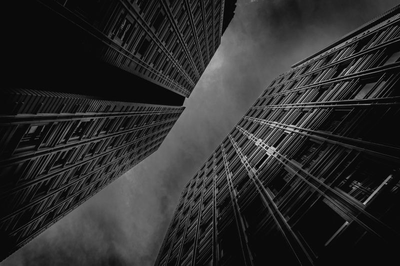 Black & white image at an unknown location in London, UK. Looking up between the two metal clad buildings, the image definitely has a Gotham City feel to it